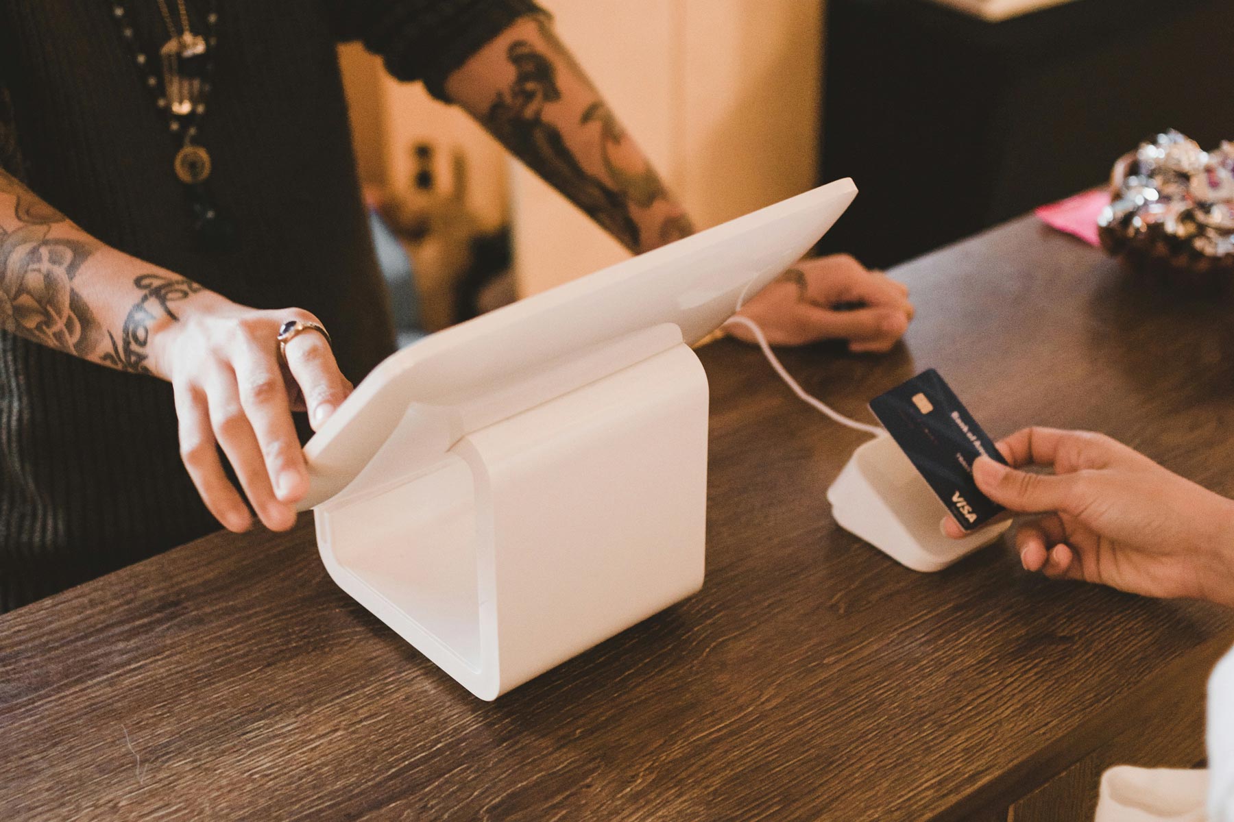 Close up of a hand tapping a payment card against a card reader which is connected to a till system. The card reader and till system are sitting on a wooden counter. Behind the counter another person is interacting with the till system' screen.