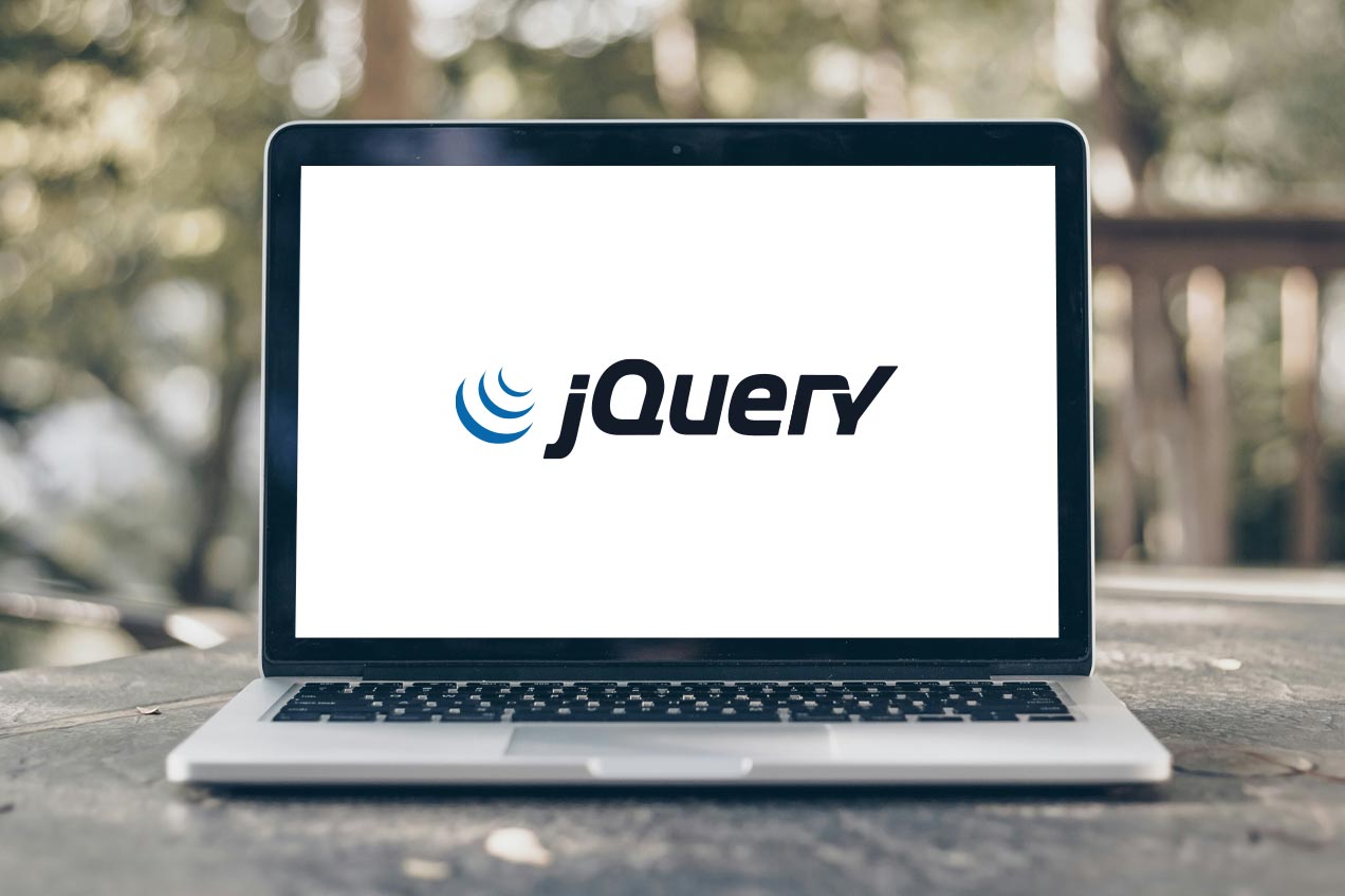 A laptop on a wooden table outdoors displays the jQuery logo on its screen, with blurred greenery in the background.