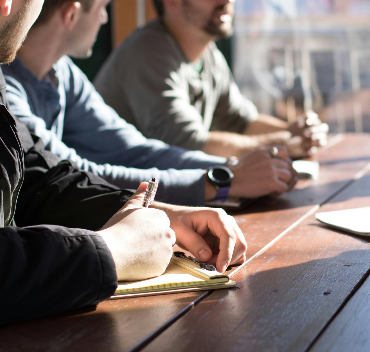 A close up of three people sitting side by side at a wooden desk. Each of them has a pen and notebook in front of them. Two of the people are looking at the third person, who looks to be talking to someone off camera on the other side of the desk.