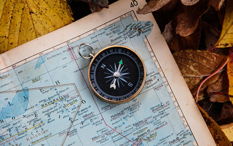 A close up of an old map laying on top of a bed of fallen leaves. On top of the map there is an old brass compass.