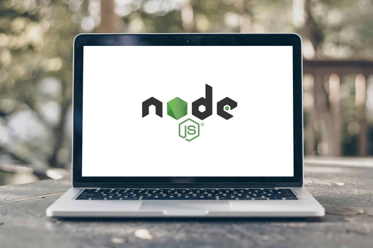 A laptop on a wooden table outdoors displays the Node JS logo on its screen, with blurred greenery in the background.