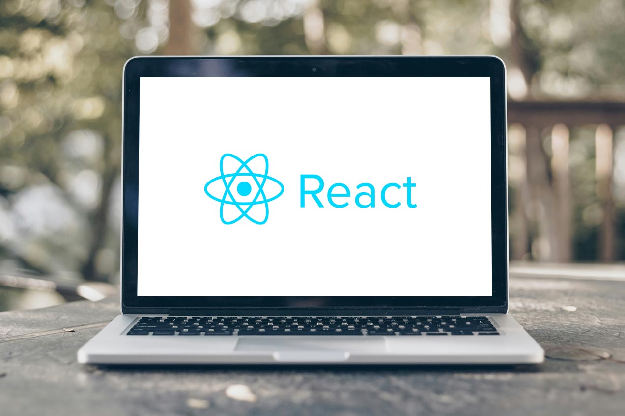 A laptop on a wooden table outdoors displays the React logo on its screen, with blurred greenery in the background.