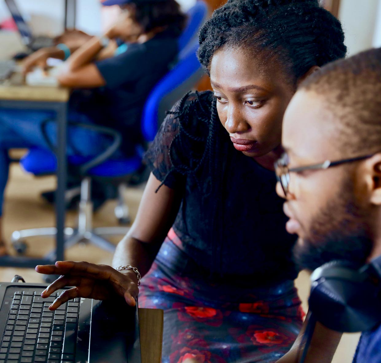 Two focused people are working together at a desk with a computer, with others in the background at a similar setting, suggesting a collaborative workspace.